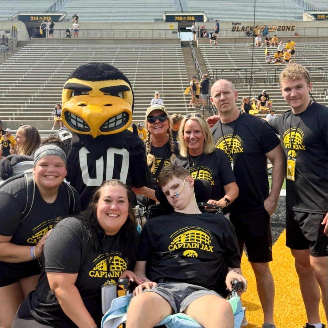 Young boy in a wheelchair, honored as a University of Iowa Kid Captain, surrounded by smiling family members all wearing matching black and yellow ‘Captain Jax’ t-shirts, posing on the field at Kinnick Stadium with the Iowa Hawkeyes mascot.